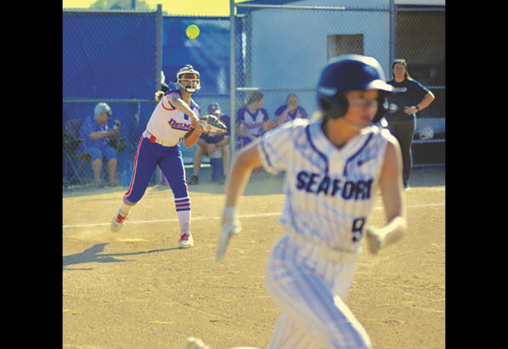 Softball game in progress: a Seaford player in a white pinstripe uniform runs toward first base, with the catcher and other players visible near home plate in the background.