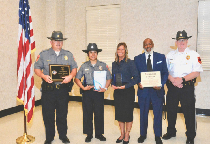 Five people standing in a room at an awards ceremony, two uniformed police officers and three civilians holding plaques and certificates beside an American flag.