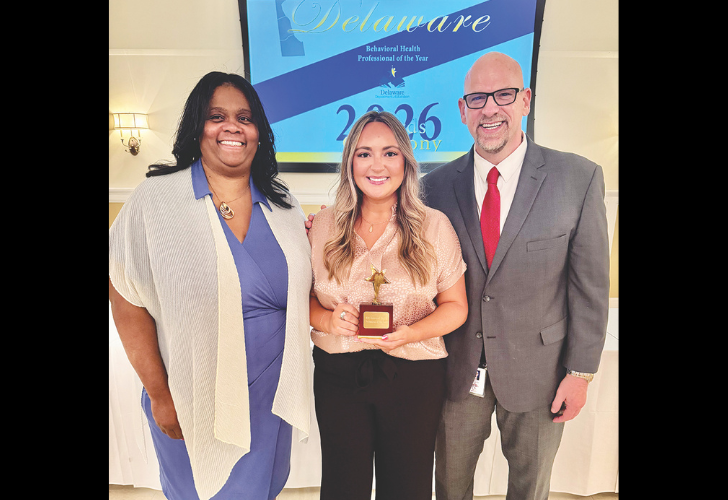Group photo of three people at an awards event; the center woman holds a trophy and smiles beside a suited man and a woman in blue attire with a Delaware banner in the background.