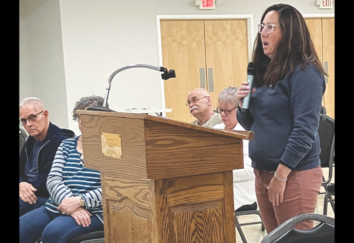 Blades Woman stands at a wooden podium holding a microphone, addressing a seated audience in a meeting room