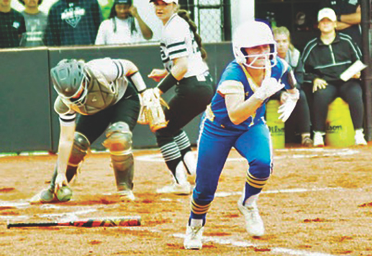 Female softball player in blue sprinting toward base with helmet on, while a catcher crouches near the base in gray protective gear.