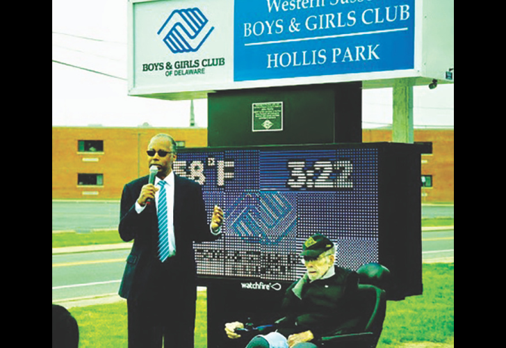 Man in suit speaks into a microphone next to a seated elderly man, outdoors at Hollis Park before a digital scoreboard and a Boys & Girls Club sign.