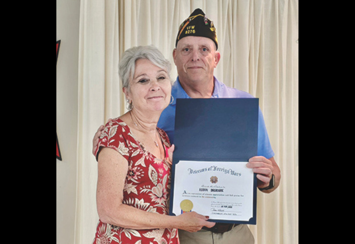 Older couple posing indoors, holding an opened certificate folder with a gold seal; the man wears a veteran’s cap while standing beside the woman in a red floral dress.