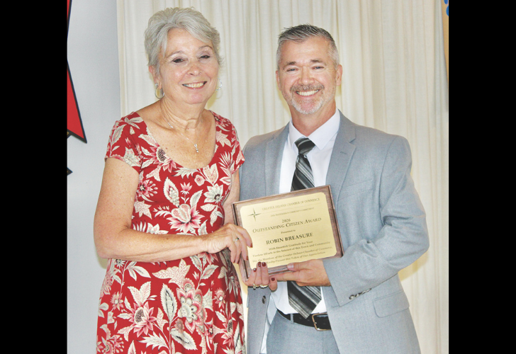 Smiling woman in a red floral dress and a man in a gray suit hold a framed award together at a ceremony.
