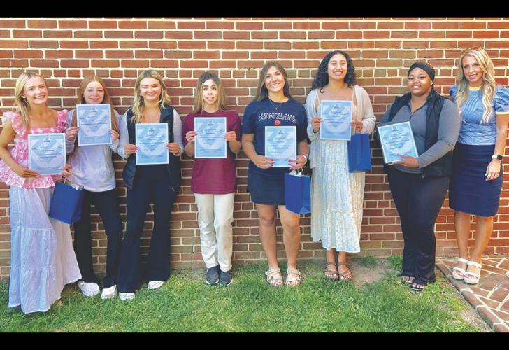 Eight young women stand in front of a brick wall, each holding a certificate of achievement and small gift bags, smiling for a group photo.