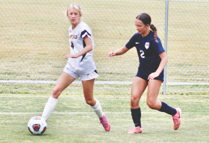Two female soccer players chase a ball on a grassy field near a chain-link fence; one in white, one in dark jersey, as the ball rolls ahead.