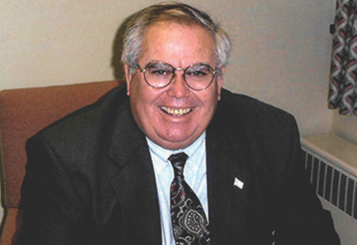 Smiling older man in a dark suit and patterned tie, wearing glasses, seated in an office setting with a window curtain in the background.