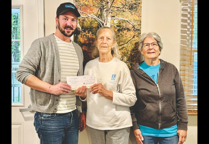 Three people indoors pose as a man hands a check to an elderly woman, with another woman smiling beside them.