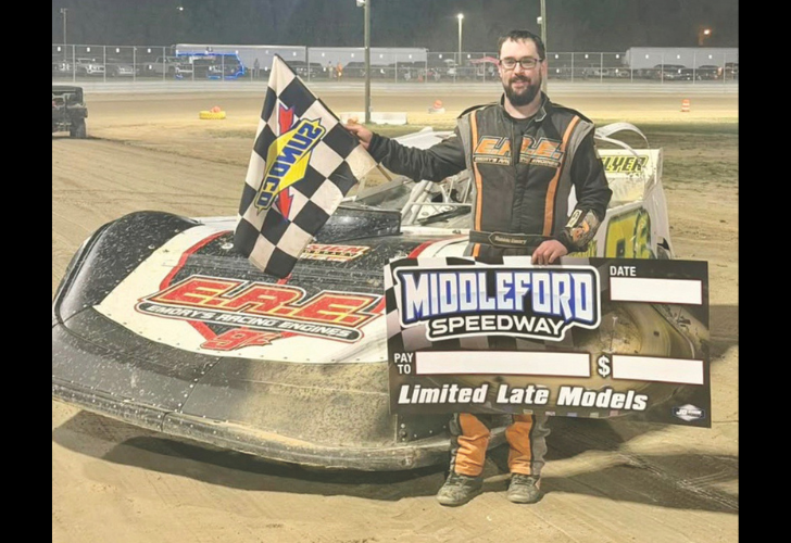 Race car driver in an orange-and-black suit stands beside a dirt-track race car at Middleford Speedway, waving a checkered flag and holding a large prize board for Limited Late Models.