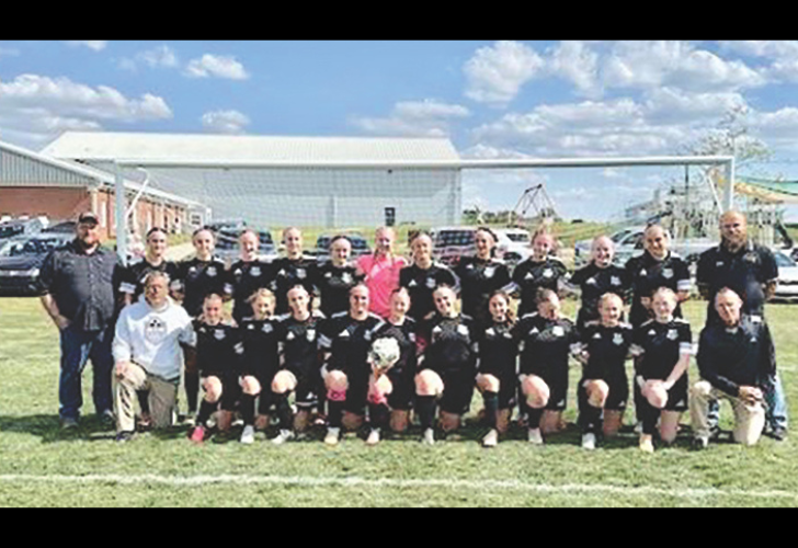Group photo of a men's soccer team in black uniforms kneeling and standing on a grass field in front of a goalpost, with coaches nearby.