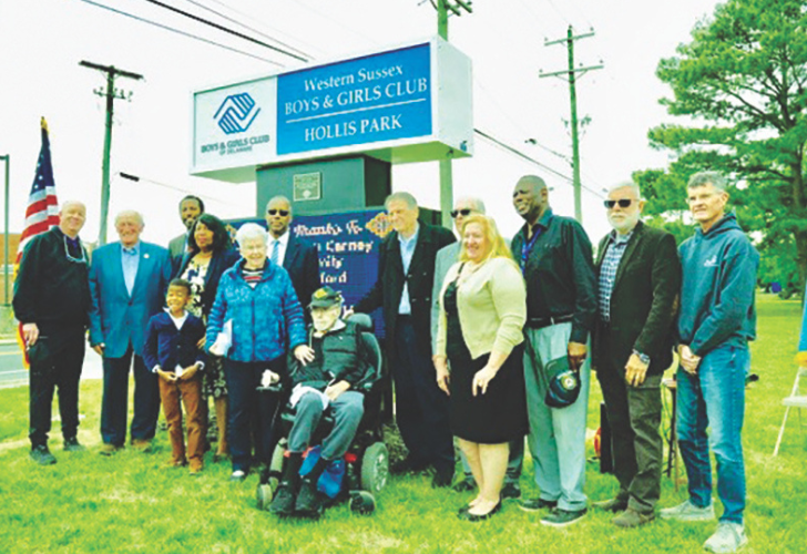 Group of diverse adults and children posing outdoors beside a sign for the Western Sussex Boys & Girls Club, Hollis Park, with a man in a wheelchair at the center.