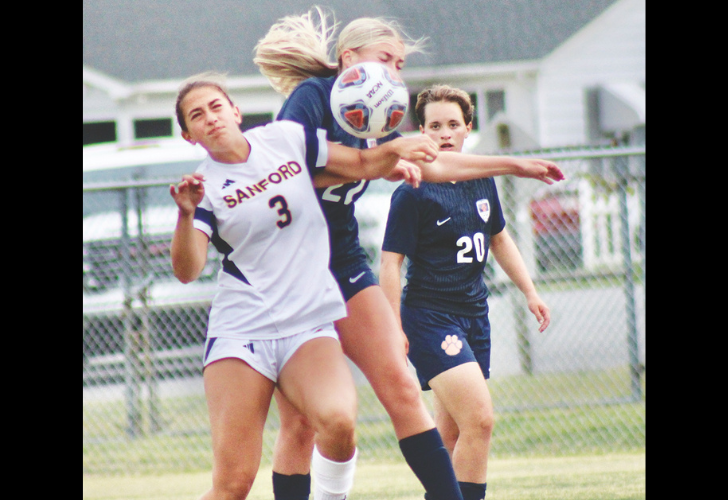 Women’s soccer players fight for the ball in midair during a match; white jersey #3 foreground, navy opponents nearby near a chain-link fence.