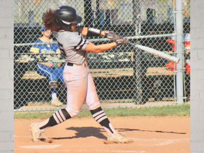 Young female softball player swinging at a pitch on a dirt field, wearing a helmet and gray uniform, with a chain-link fence in the background.