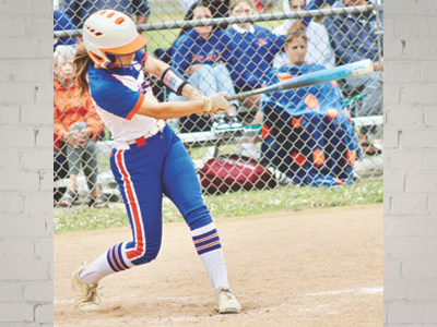 Female softball player in blue and orange uniform swings a bat during a game on a dirt field, behind a chain-link fence.