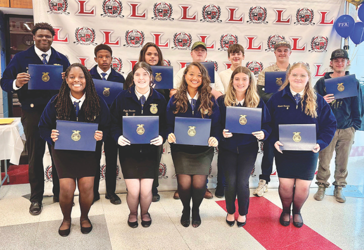 Group of students in blue FFA jackets pose with blue award folders at a ceremony in front of a logo backdrop.