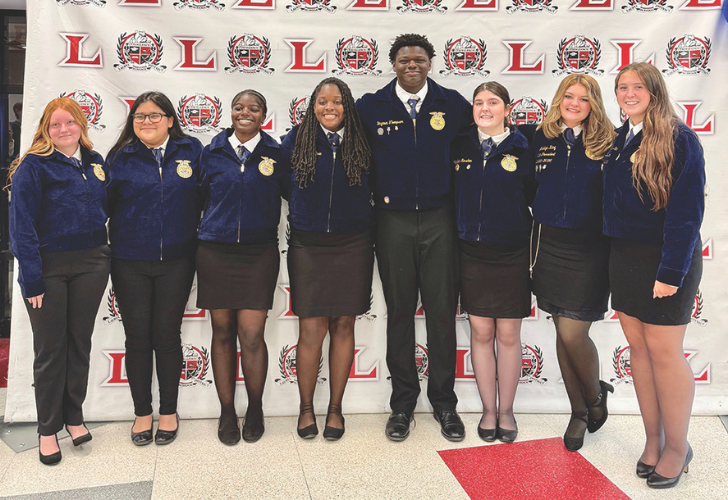 Nine students in matching navy jackets and dark skirts or pants pose for a group photo in front of a step-and-repeat backdrop featuring the school logo.
