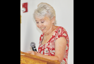 Woman with short gray hair speaking into a microphone at a lectern, wearing a red floral blouse and smiling