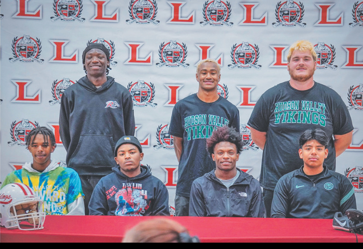 LAU Signing L27 Seven young men pose for a signing-day photo in front of a logo-covered backdrop; several wear Hudson Valley Vikings shirts, with a football helmet on the table.
