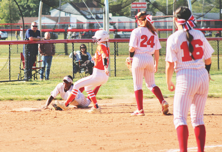 Louis L25 Softball game in progress; a runner slides into a base as teammates and spectators watch behind the fence on the grassy field.