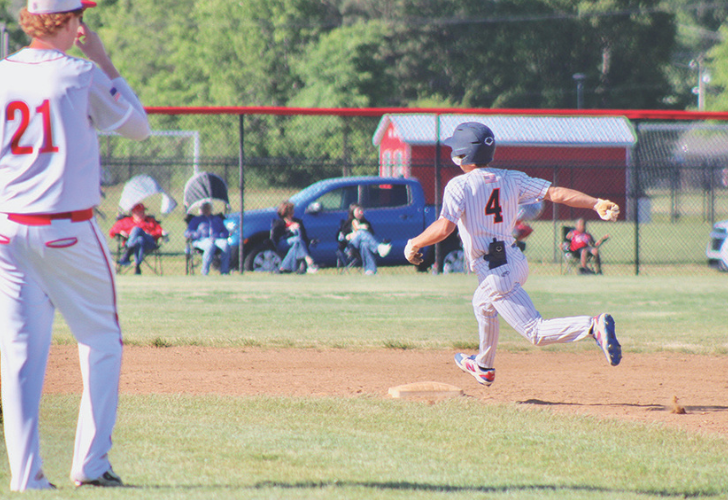 McCabe L25 Youth baseball scene: a runner in gray pinstripes slides toward first base as a fielder in white with red 21 watches nearby.
