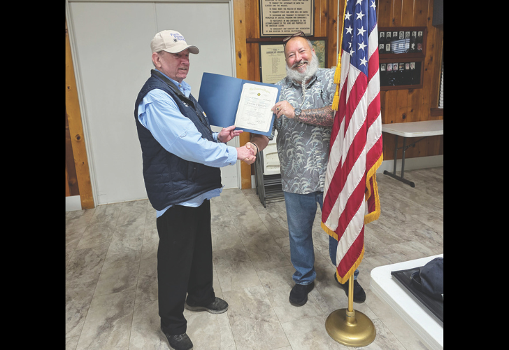 Two men shake hands as one receives a certificate in a room with wood paneling and an American flag nearby.