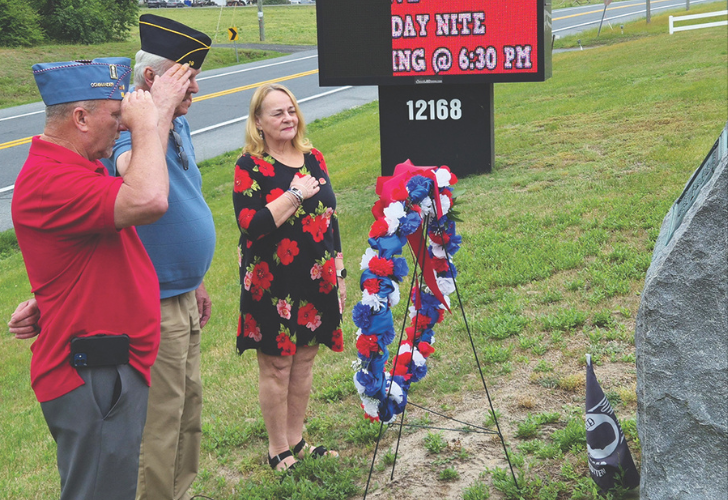 Three people, two veterans and a woman in a floral dress, stand by a patriotic wreath at a roadside memorial, saluting or paying respects near a stone marker and digital board behind them.