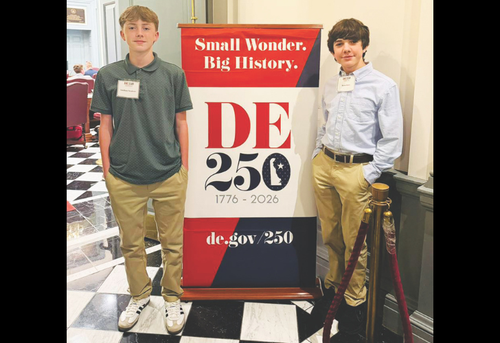Two boys stand beside a DE 250 banner reading Small Wonder. Big History. 1776-2026, at a formal event.