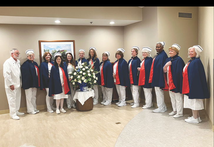 Nurse Honor Guard Group of people in blue capes with red lining and white hats standing in a hallway around a large flower arrangement on a stand.