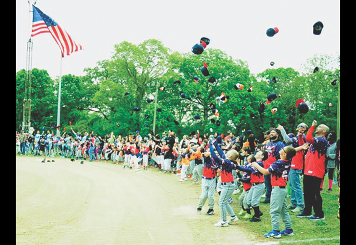 Crowd of kids and adults in red vests cheer and toss hats in the air beside a dirt path and green trees, with an American flag visible. (celebration)
