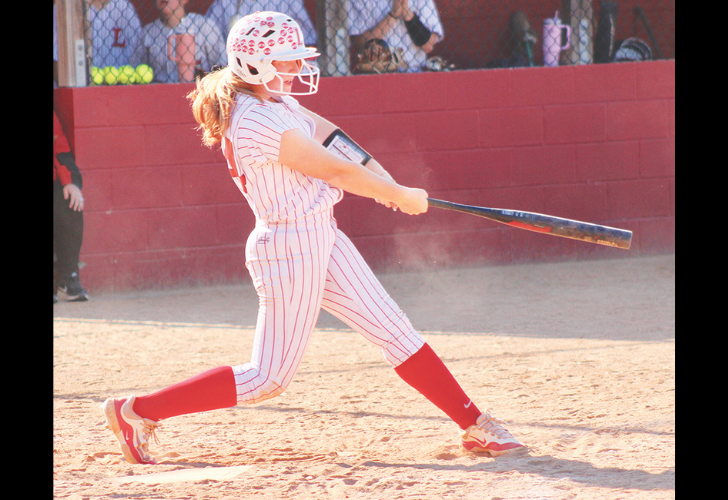 Softball batter in a white pinstripe uniform and red socks swings a bat on a dirt field with a red wall behind her.