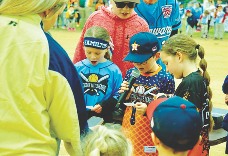 Group of young children at a youth baseball event, some wearing helmets and caps, one holding a microphone while a coach oversees a activity.