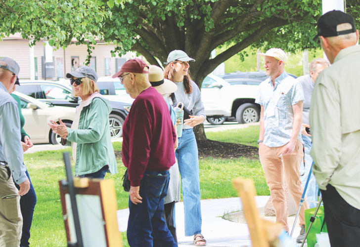 Group of adults standing and chatting in a park-like outdoor setting with trees and parked cars in the background.