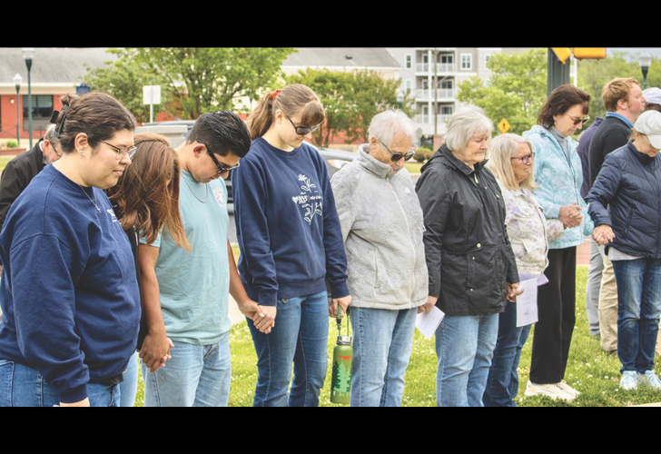 Group of people of various ages standing in a row on a grassy area, some holding hands and papers, in a moment of silence at an outdoor gathering.
