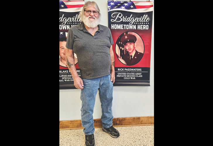 Rick Passwaters Older man with gray beard and glasses stands in front of patriotic posters featuring a soldier portrait and 'Bridgeville Hometown Hero' banners, indoors.