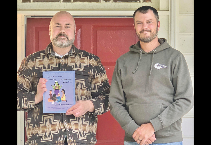 Two men stand side by side in front of a red door; the man on the left holds a purple cartoon book toward the camera.