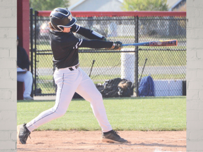 Baseball batter in a dark jersey and white pants swings a bat during a game on an infield with a chain-link fence in the background.