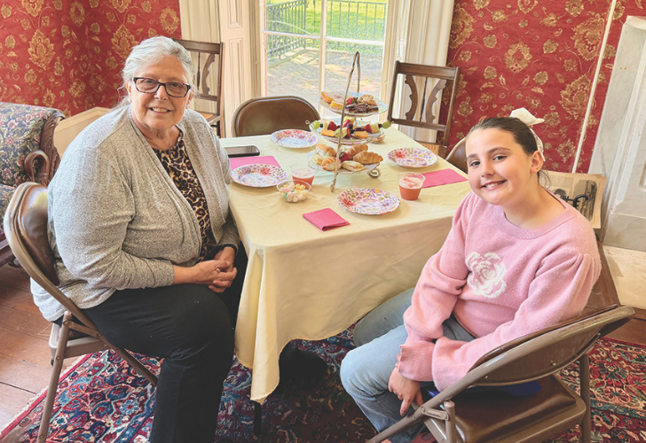 Two generations smiling at a tea party for two, seated at a round table with a tiered tray and pink decor in a sunlit dining room.