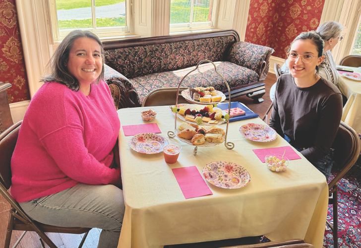 Two women smiling at a tea party, seated at a table with a two-tier pastry stand and pink napkins.
