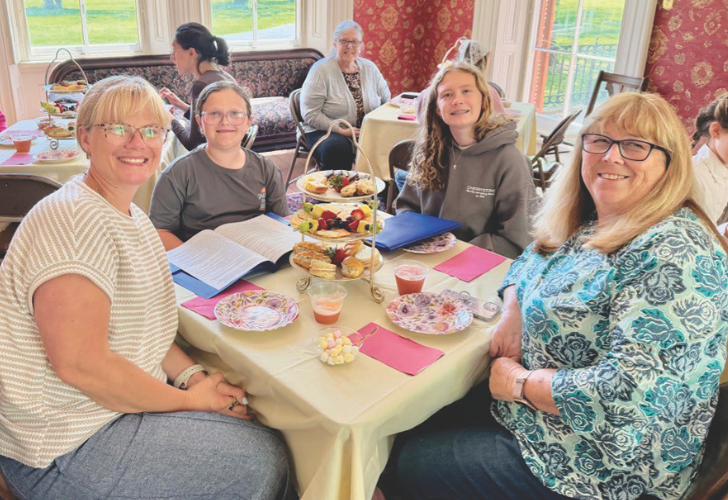 Group of women seated around a table with tiered snacks, enjoying tea and conversation in a bright room.