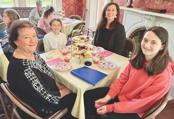 Group of four generations enjoying a tea party at a warmly decorated table with pastries and pink napkins on the tablecloth.
