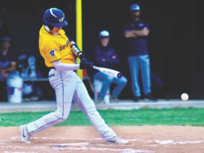 Young baseball player in a yellow jersey and blue helmet swings at a pitch on a dirt field, ball near the bat as it approaches home plate.