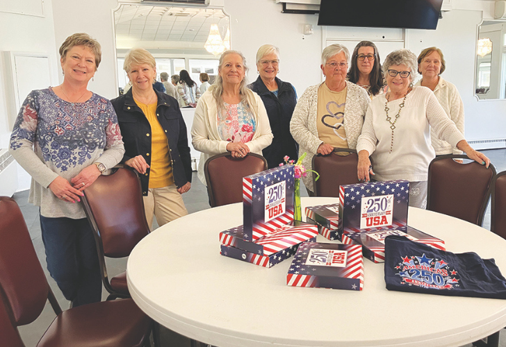 Group of eight women standing behind a round table with USA-flag themed gift boxes for a 250th celebration.