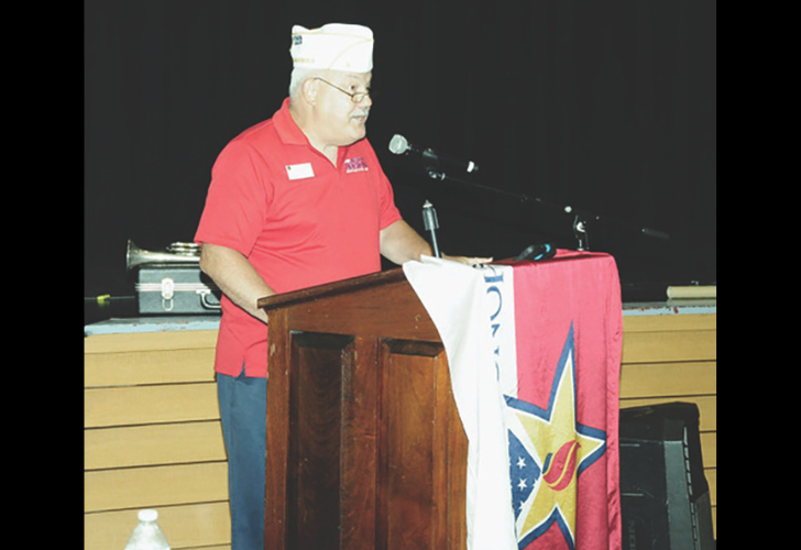 Man in a red polo shirt and white cap speaks at a wooden podium on a stage, microphone in front, star-banner draped over the podium