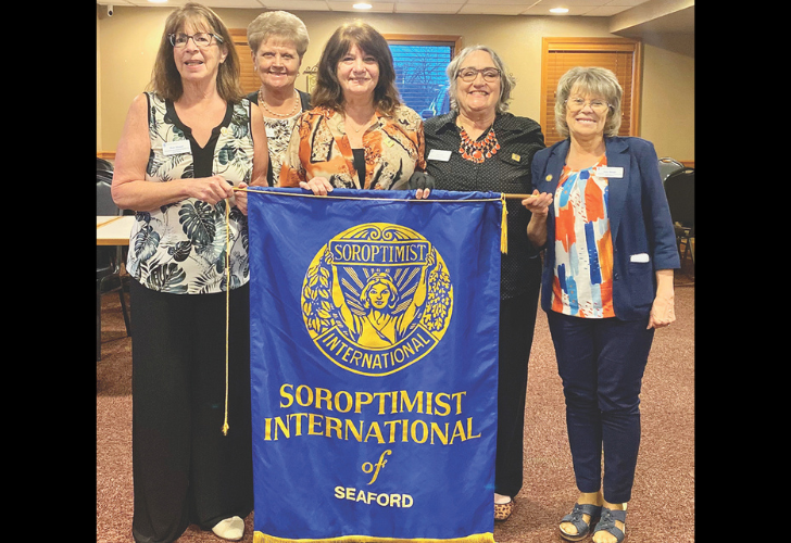 Five women stand together in a meeting room, holding a blue Soroptimist International banner reading Seaford.