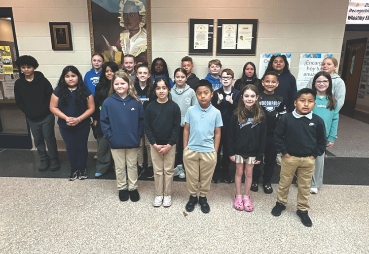 Diverse group of about 20 students posing for a class photo in a school hallway with framed certificates on the wall behind them.