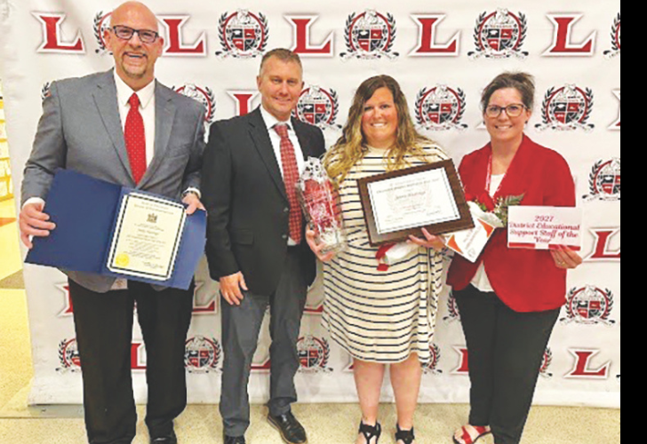 Group of four adults at an awards event; two hold plaques/trophies and a framed certificate, backdrop features red L logos.