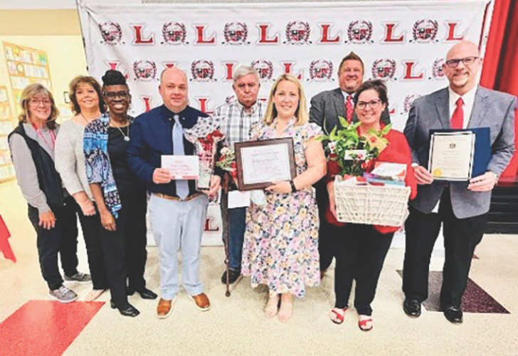 Nine adults posing at an awards ceremony; center woman in a floral dress holds a framed certificate, another woman carries a gift basket, others hold awards.