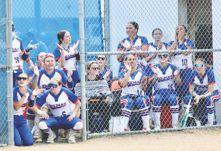 Del Mar softball team cheering and clapping behind a chain-link fence, players in white-blue uniforms with numbers, dugout area in the background.