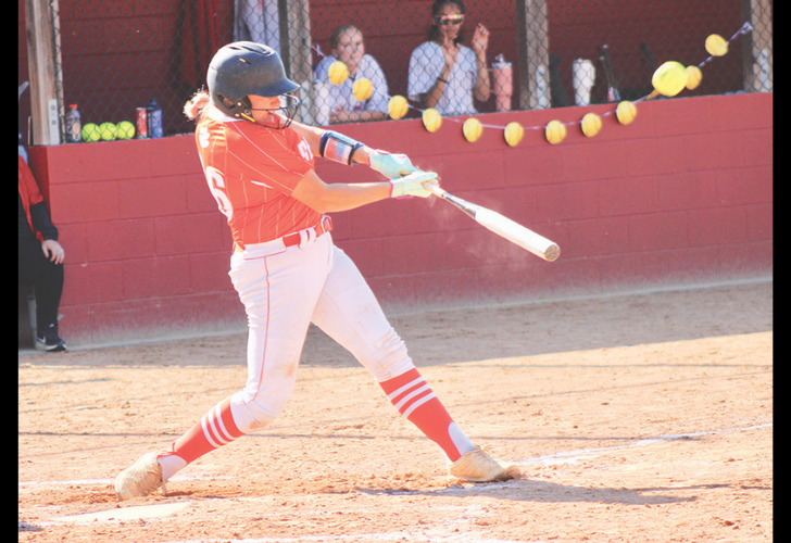 Willey L25 Female softball player in a red uniform swings a bat at a pitched ball during a game, with spectators behind a chain-link fence.