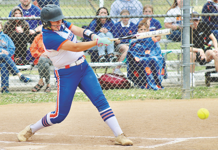 Female softball player in blue and white prepares to hit, swing connecting with a pitched ball during a game behind a chain-link fence.